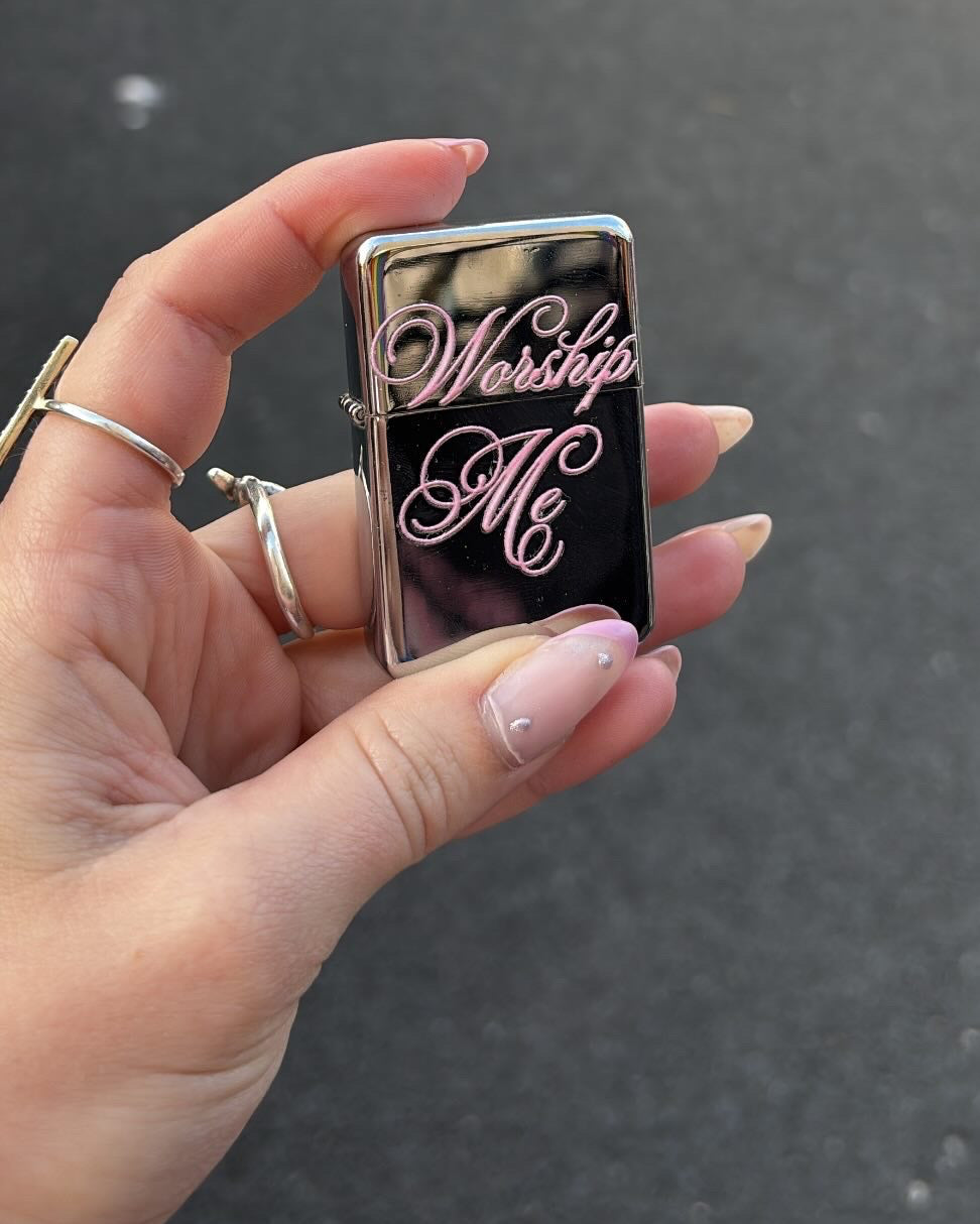 A hand holding a silver Zippo-style lighter with a black center panel and pink script text reading 'Worship Me'. The background is a blurred outdoor street scene with market stalls, and a white text bubble at the top says 'WORSHIP ME LIGHTER!'.